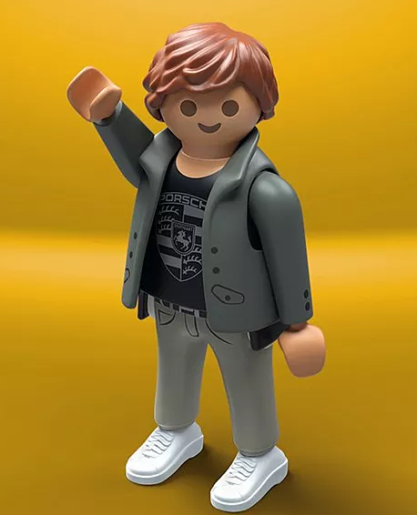 Eine Playmobil-Figur mit braunen Haaren und einer grauen Jacke, trägt ein schwarzes Shirt mit Porsche-Logo und weiße Turnschuhe, auf gelbem Hintergrund.