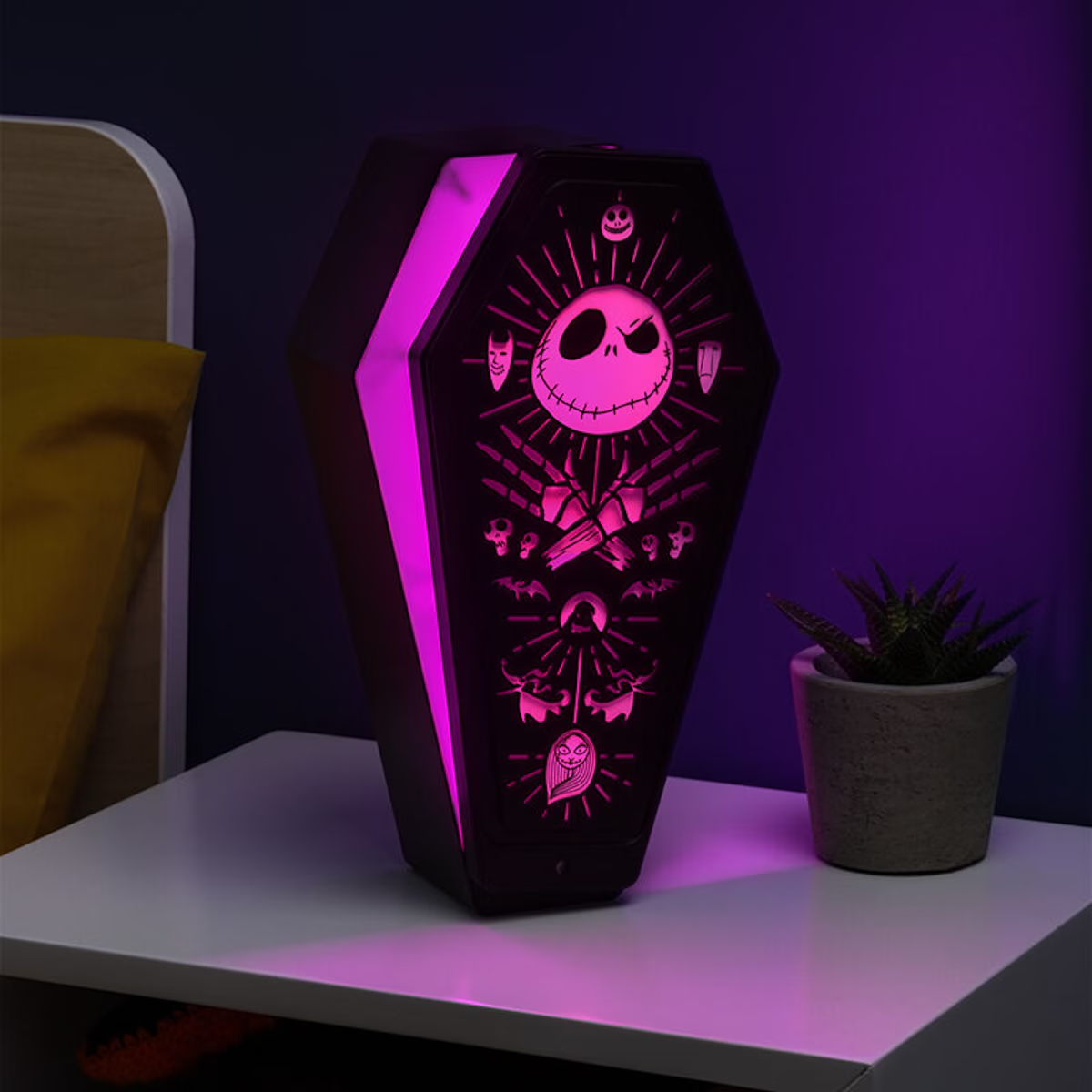 Una lámpara con forma de ataúd con diseño de Jack Skellington, que emite luz rosa, está sobre una mesa blanca. Una planta a la derecha.