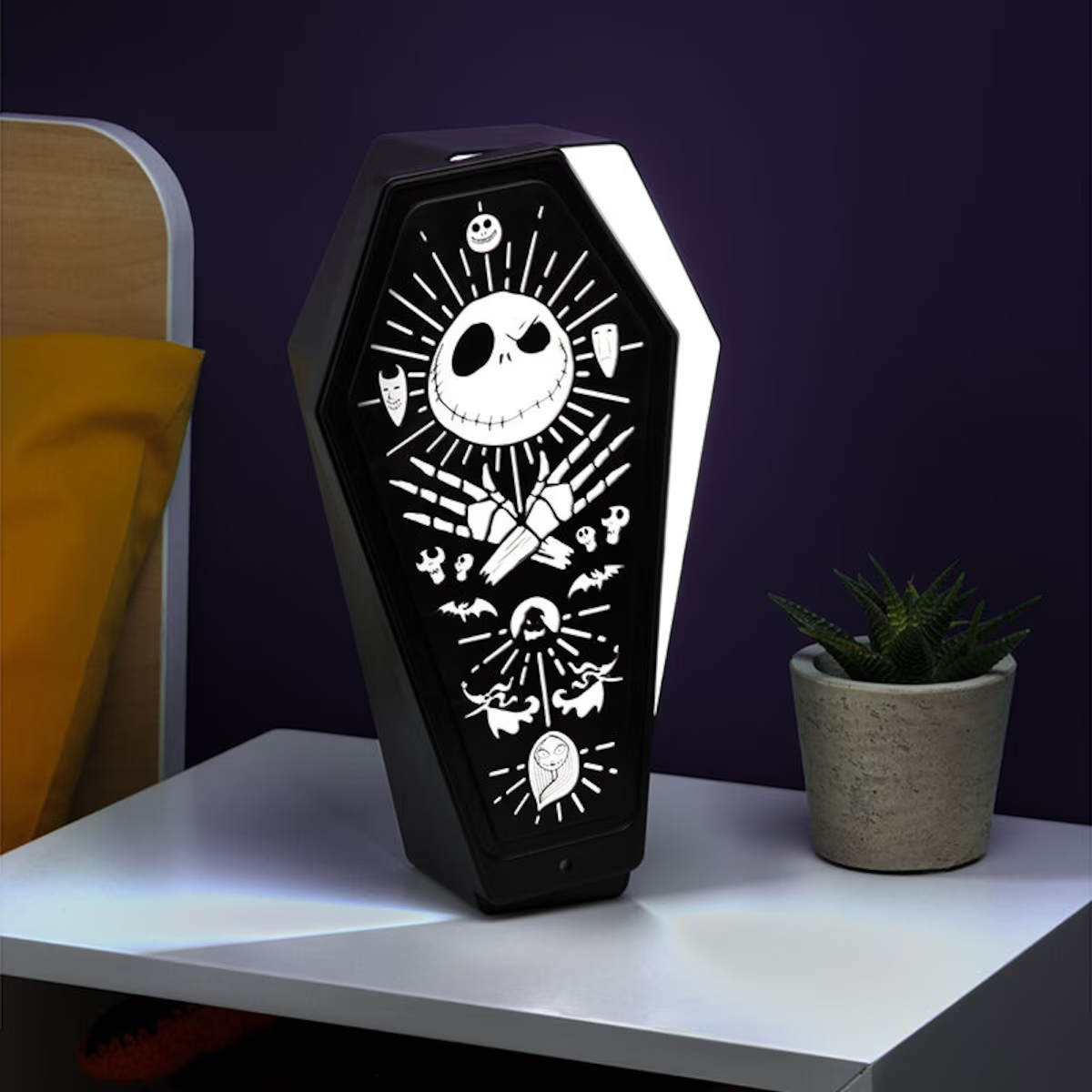 Una lámpara con forma de ataúd con diseño de Jack Skellington está sobre una mesa blanca. Una pequeña planta en maceta está al lado.
