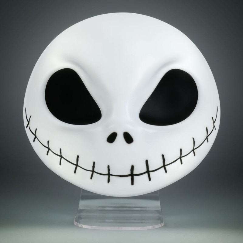 Una máscara blanca de Jack Skellington con ojos, nariz y sonrisa cosida negros. Se encuentra sobre un soporte transparente contra un fondo gris.