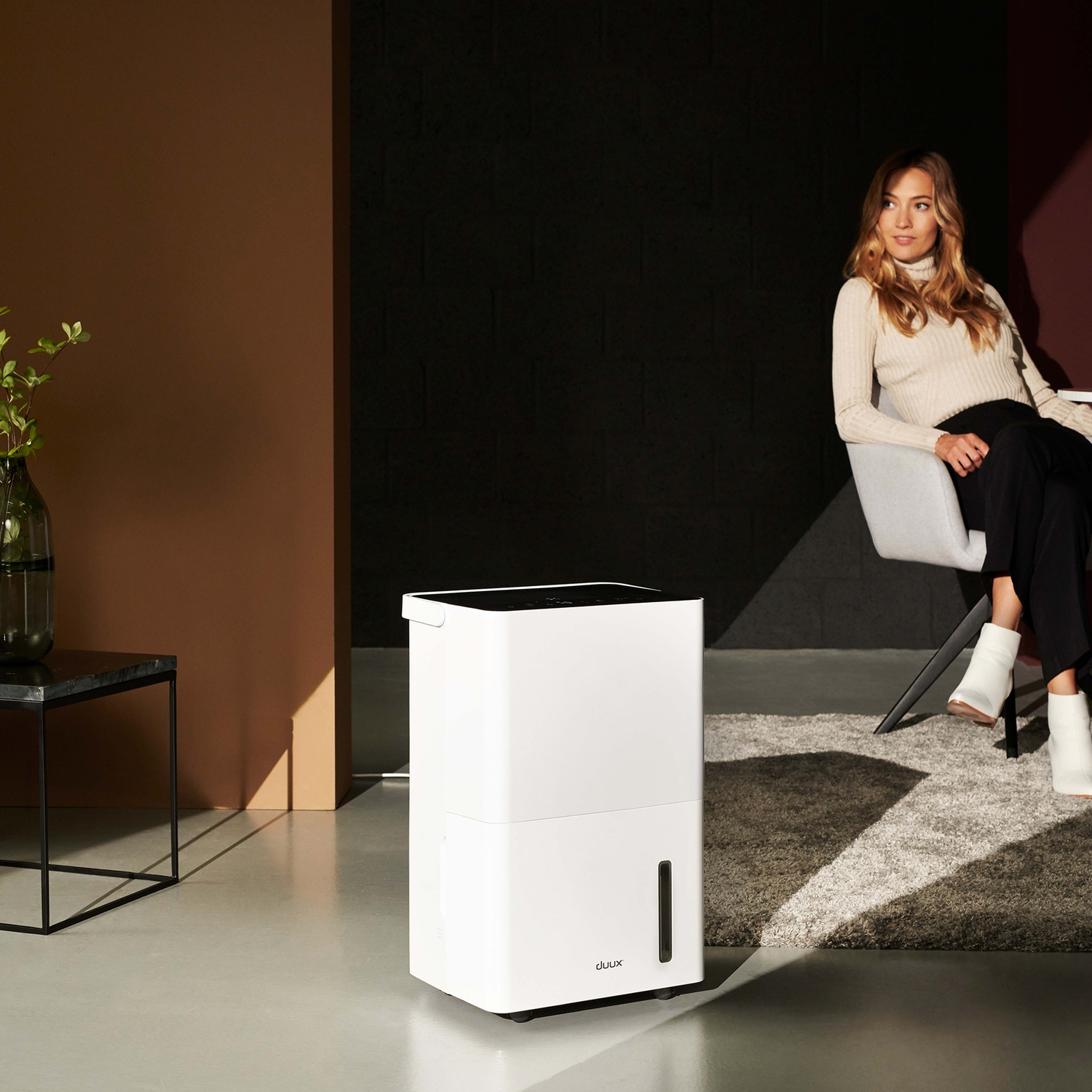 A woman sits near a dehumidifier. The room has brown and black walls. A plant is on a table. The floor is light grey.