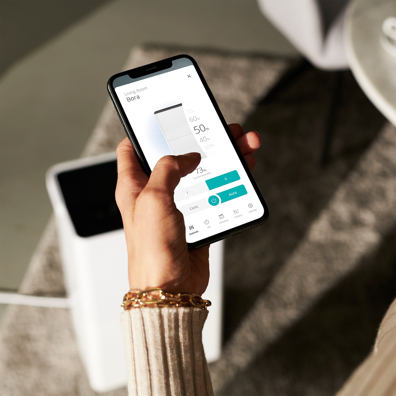 A hand holding a smartphone showing a smart home interface. The phone displays humidity levels. A gold bracelet is visible.