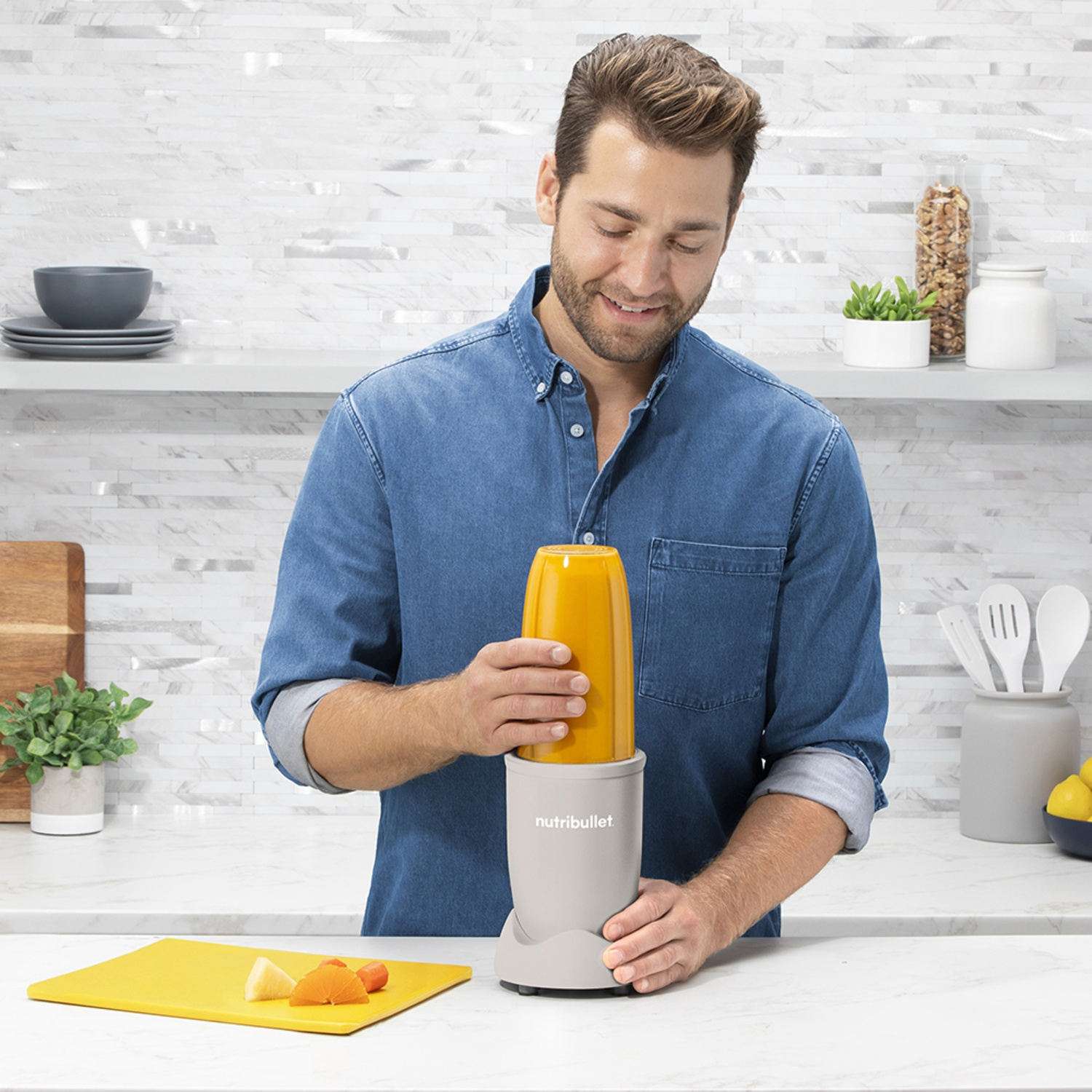 A man in a blue shirt is assembling a Nutribullet. A yellow cutting board with fruit sits nearby. Kitchen background.