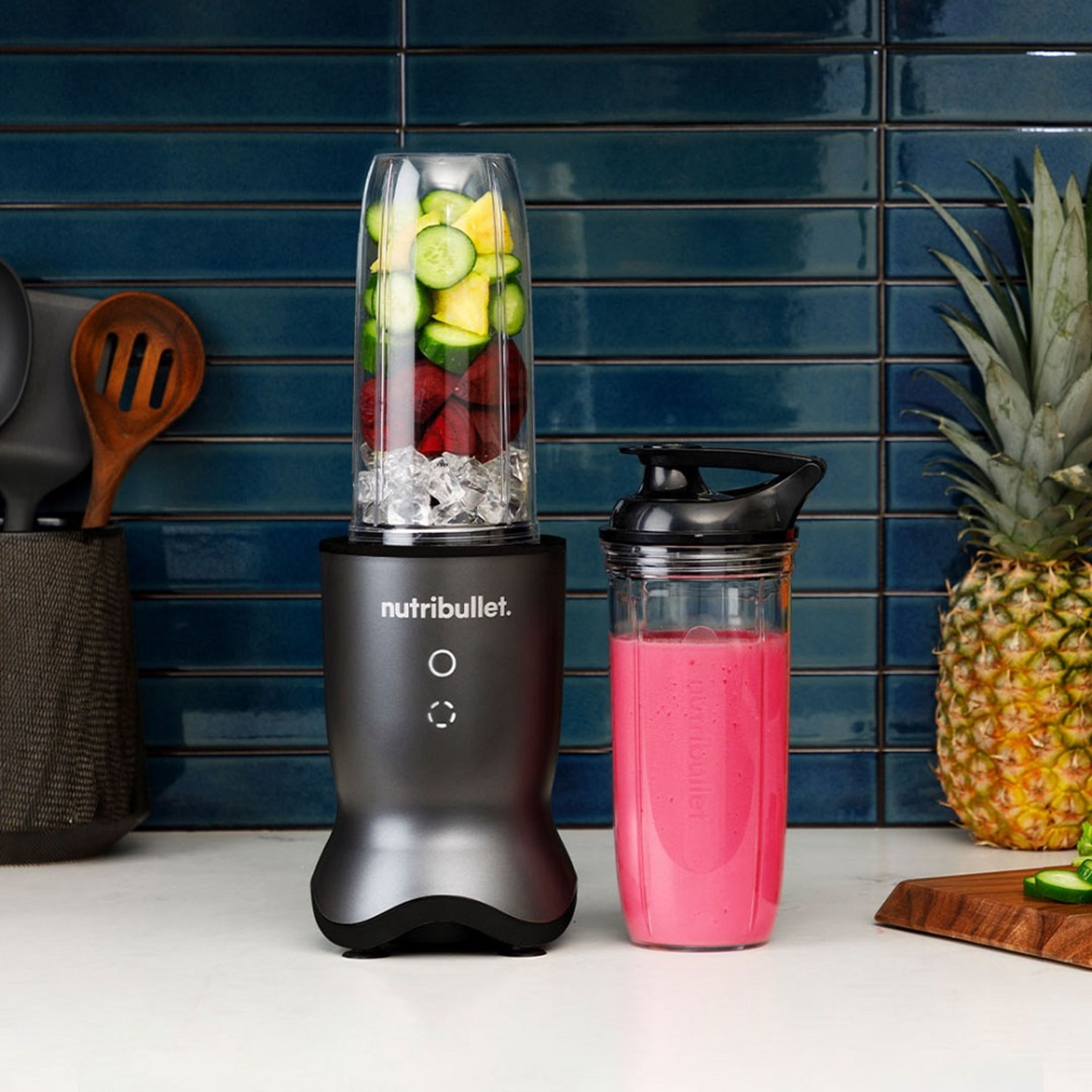 A Nutribullet blender with ingredients like cucumber and pineapple. A pink smoothie cup is next to it. A pineapple is on the right.