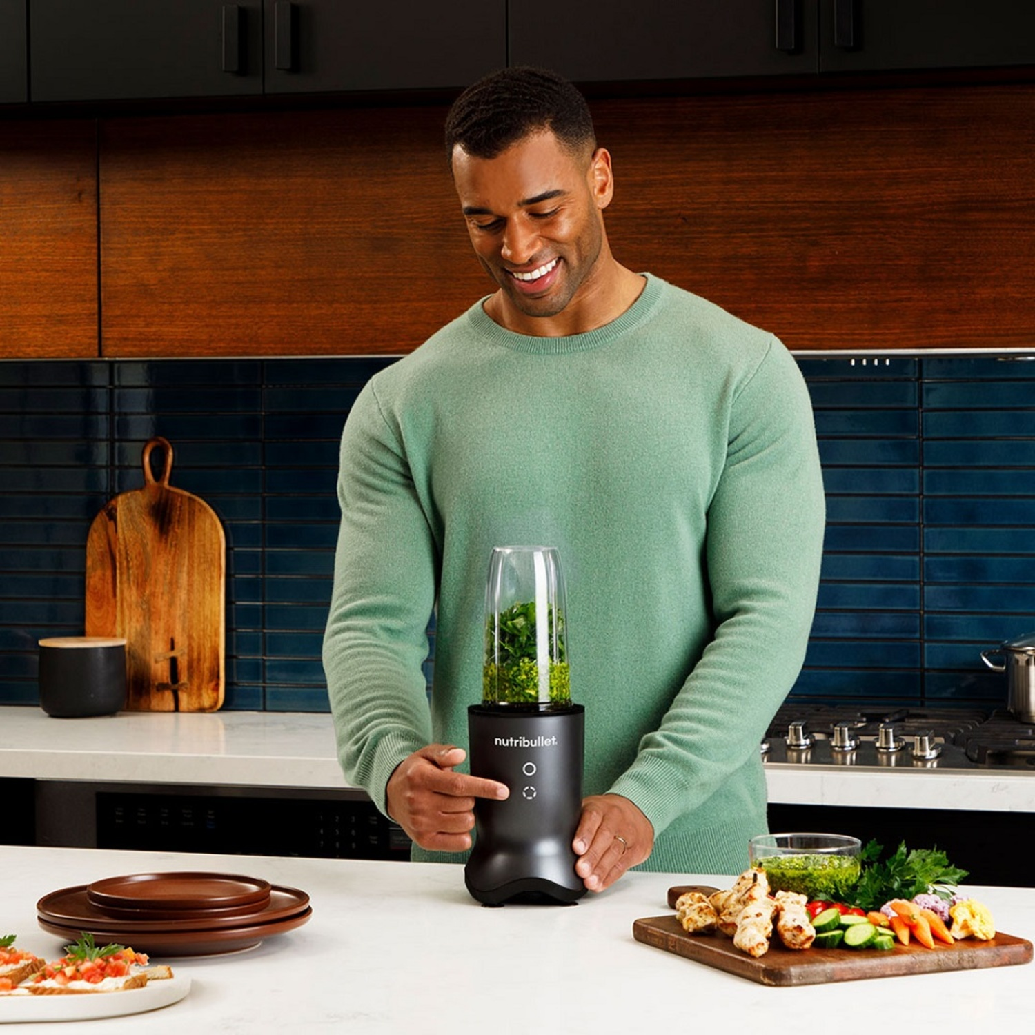Man using a Nutribullet blender. He smiles at the device filled with green ingredients. Food is on the counter.