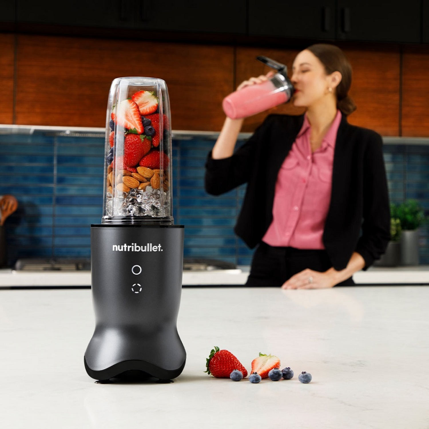 A woman drinks a smoothie. A blender with fruit stands on a counter. Strawberries and blueberries are in front of it.