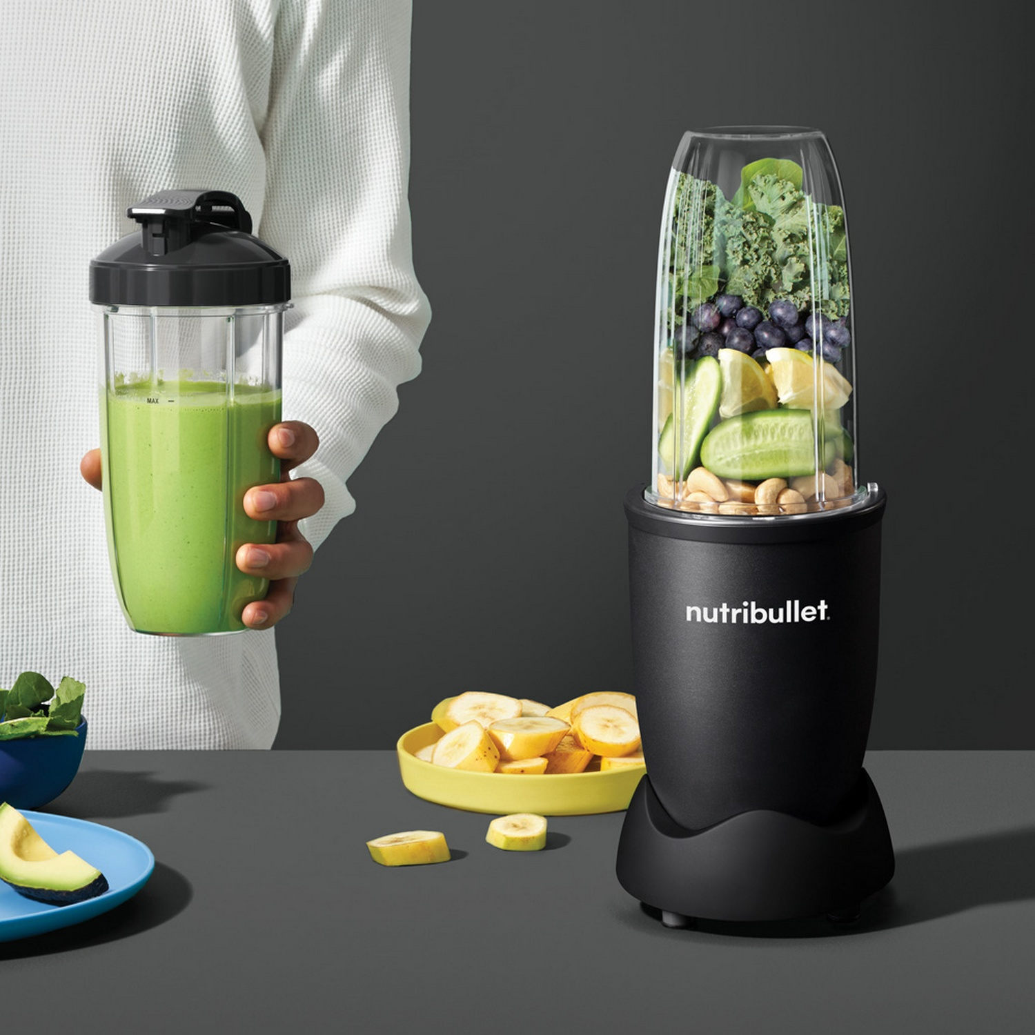 Een persoon houdt een groene smoothie vast. Een blender is gevuld met fruit en groenten. Bananen, avocado en spinazie staan ook op tafel.