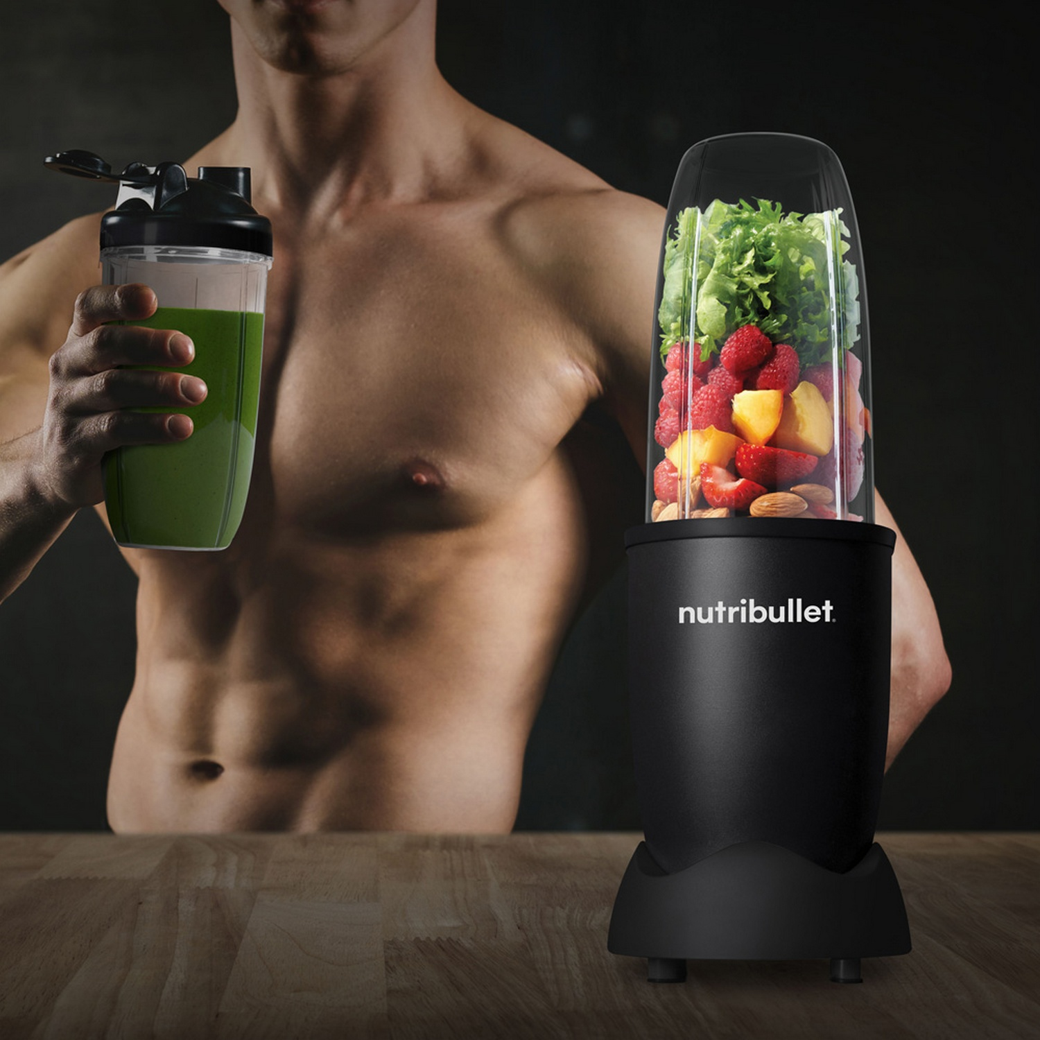 Een man houdt een groene smoothie en een Nutribullet vol fruit en groenten vast. De achtergrond is donker.