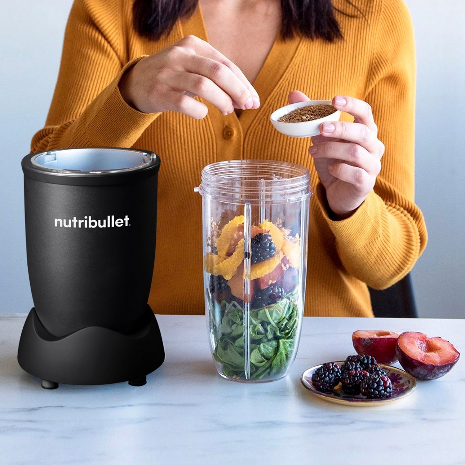 Een persoon voegt zaden toe aan een blender gevuld met fruit en spinazie. Een Nutribullet staat links. Pruimen en bessen staan rechts.