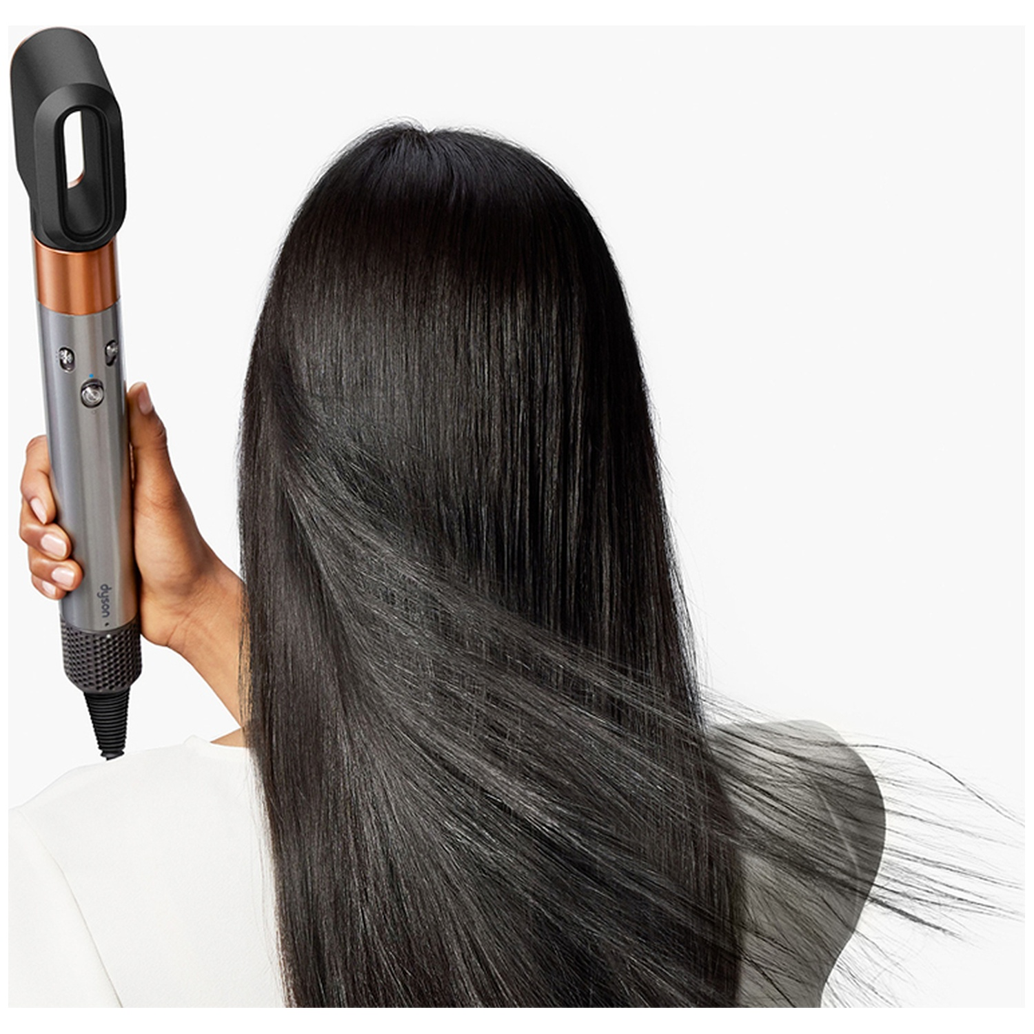 A person holding a hair dryer, facing away. Long, black hair flows. The dryer is silver and black, with a copper accent.