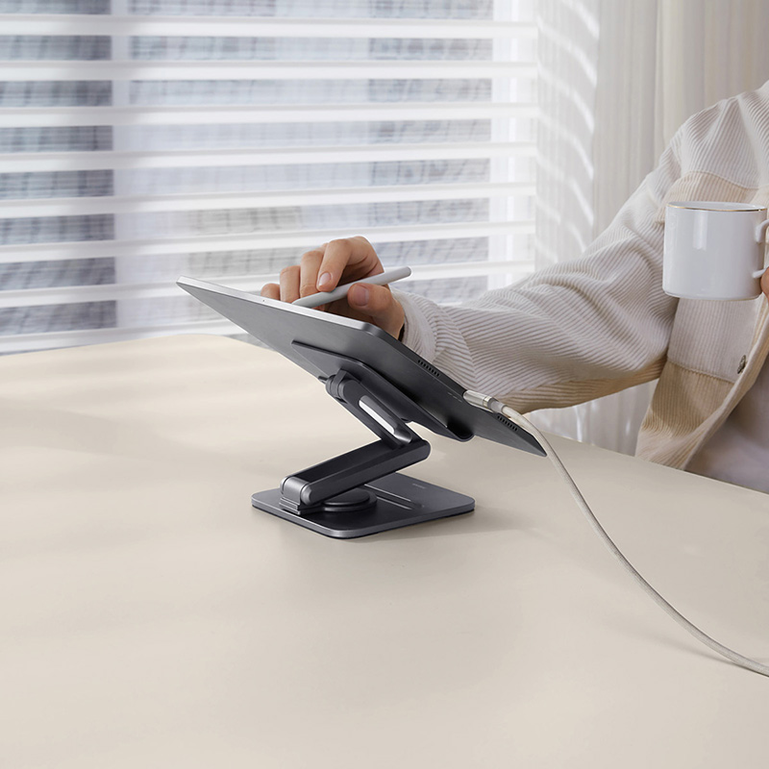 Una persona usa una tableta con un lápiz óptico, sosteniendo una taza blanca. Tableta en un soporte, cable blanco. Persiana en el fondo.