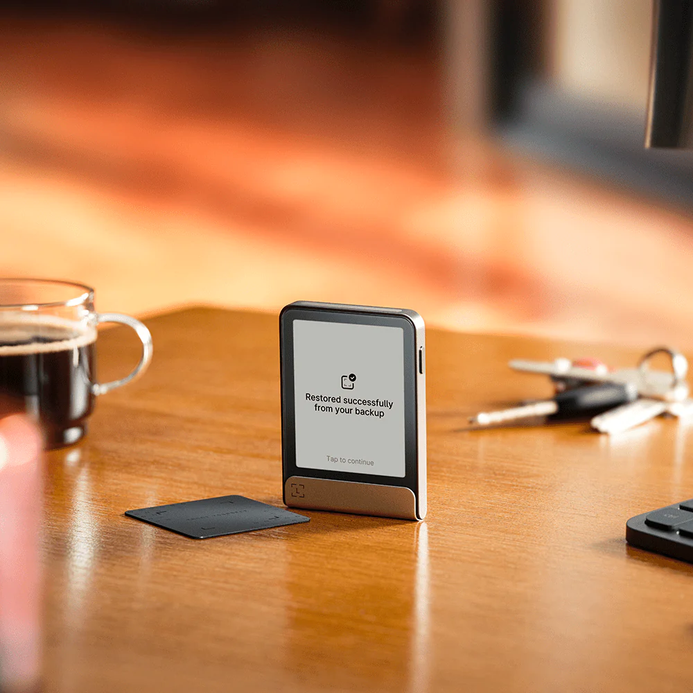 Ein telefonähnliches Gerät mit Bildschirm zeigt 'Erfolgreich wiederhergestellt'. Schlüssel, eine Kaffeetasse und eine Karte liegen auf einem Holztisch.