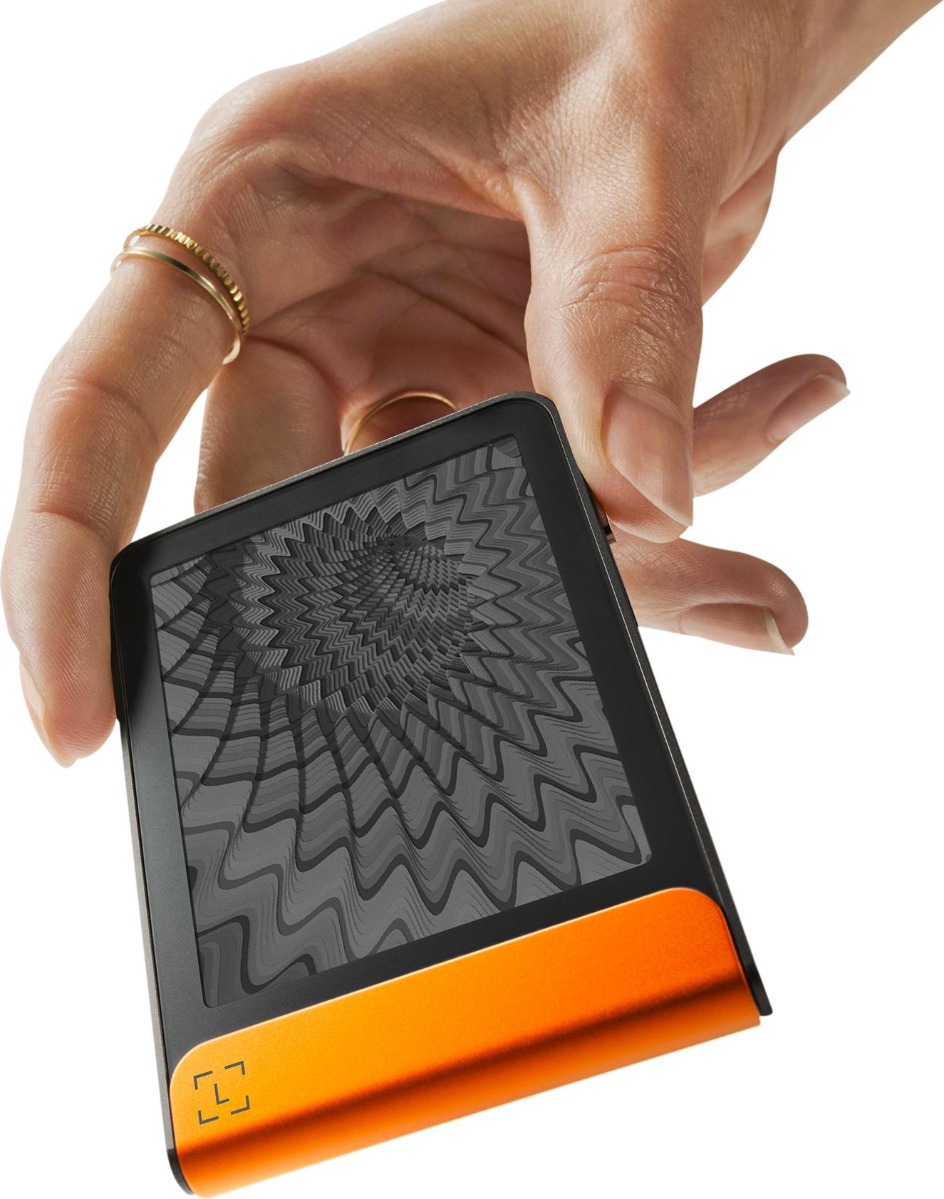 Une main tient un appareil noir et orange avec un écran à motifs. La main porte des bagues dorées. Fond blanc.