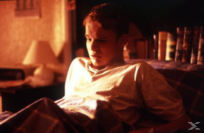 Ein junger Mann sitzt besorgt auf einem Bett. Eine Lampe und ein Bücherregal befinden sich im Hintergrund, beleuchtet von warmem Licht.