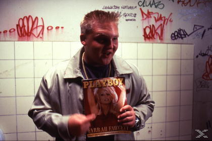 Ein Mann hält ein Playboy-Magazin mit Farrah Fawcett auf dem Cover. Er steht in einem gefliesten Raum mit Graffiti an der Wand.