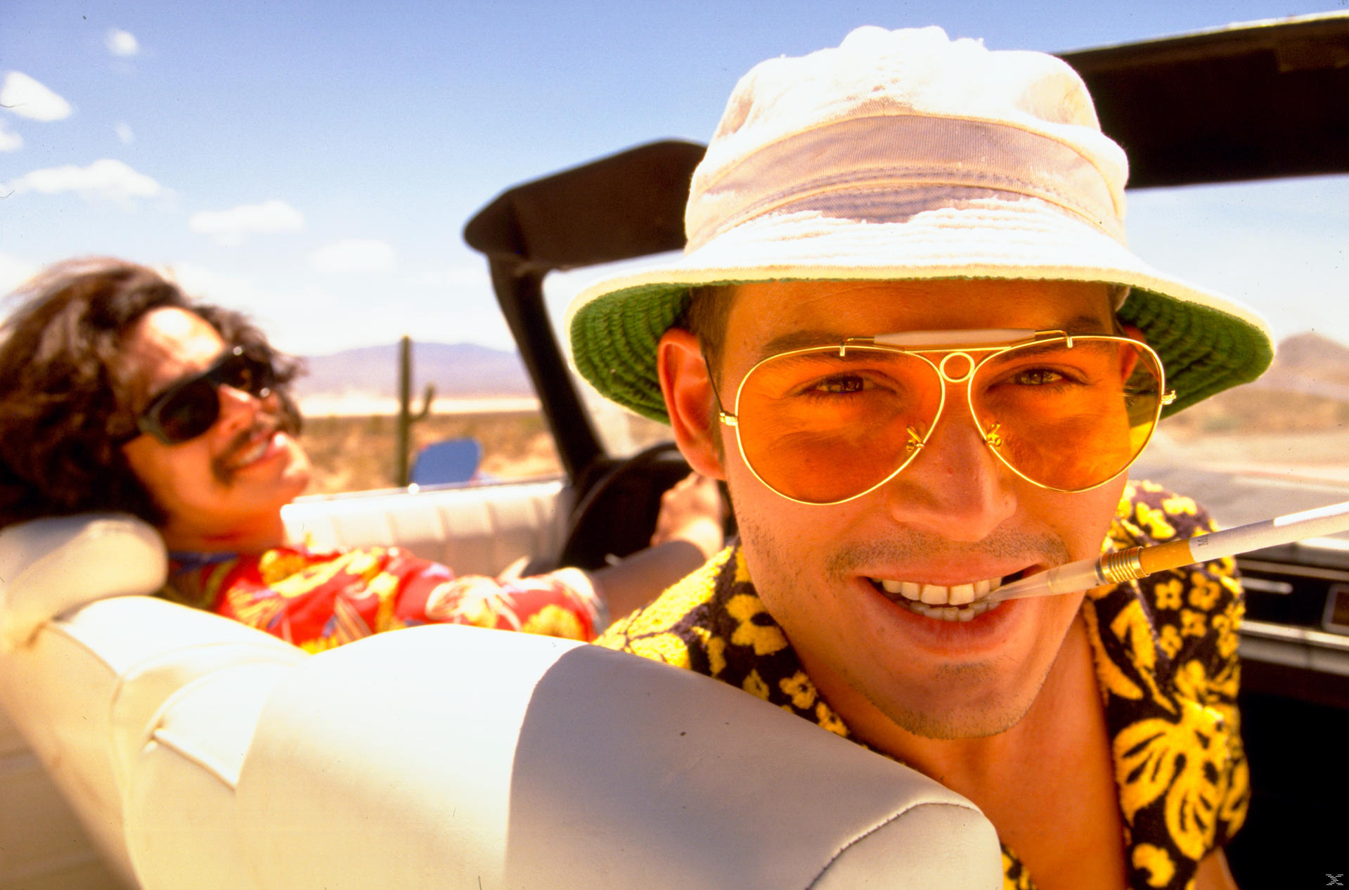 Mann mit Sonnenbrille und Hut, Zigarette im Mund, lächelt im Auto. Eine weitere Person im Hintergrund.