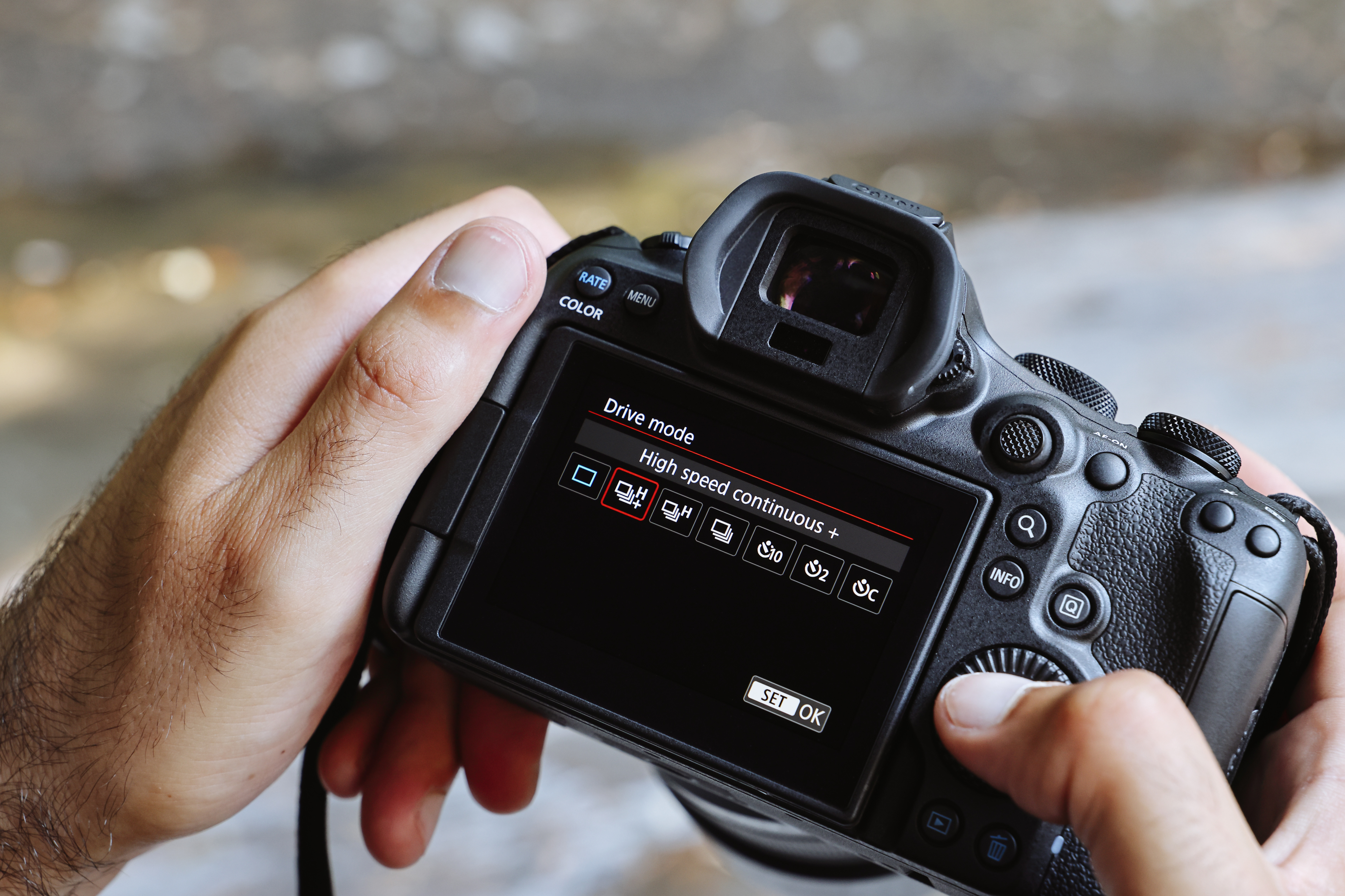 Eine Person hält eine schwarze Kamera, die 'Drive Mode'-Optionen anzeigt, mit einem Finger, der eine Taste drückt. Der Hintergrund ist unscharf.
