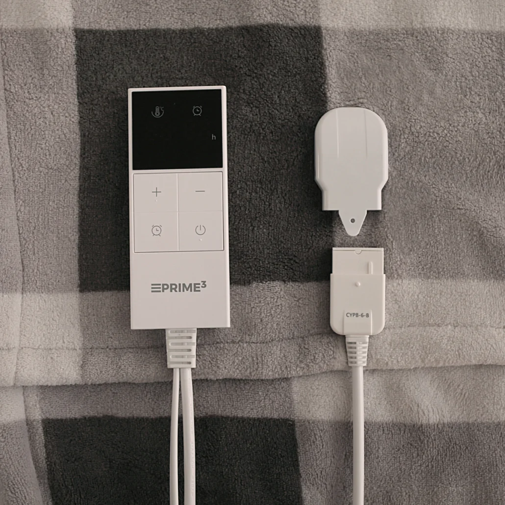 Biały kontroler ePrime3 i adapter znajdują się na szarej kraciastej kołdrze. Kontroler ma przyciski i czarny wyświetlacz.