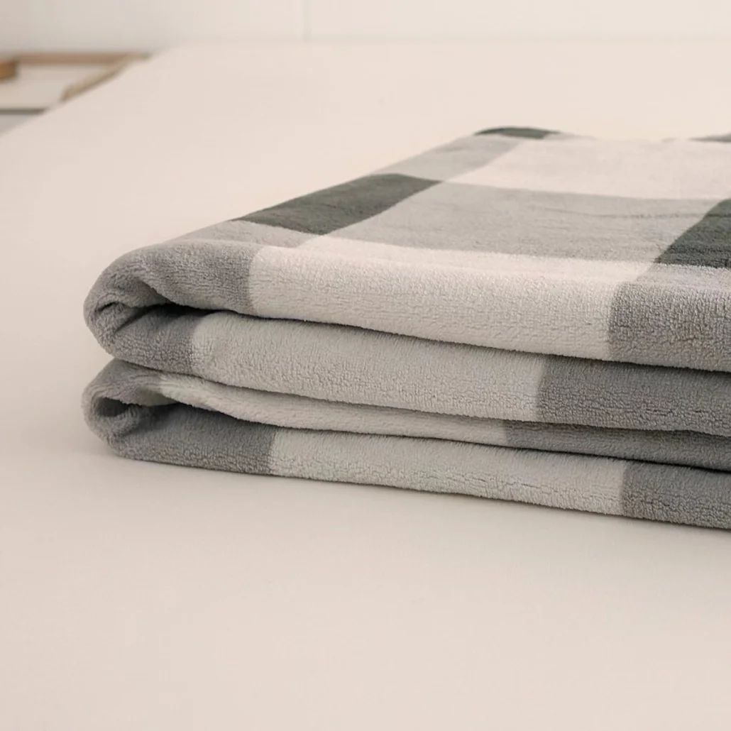 Złożony koc z szaro-białym wzorem w kratę leży na białej powierzchni. Koc jest miękki, a zagięcia widoczne.