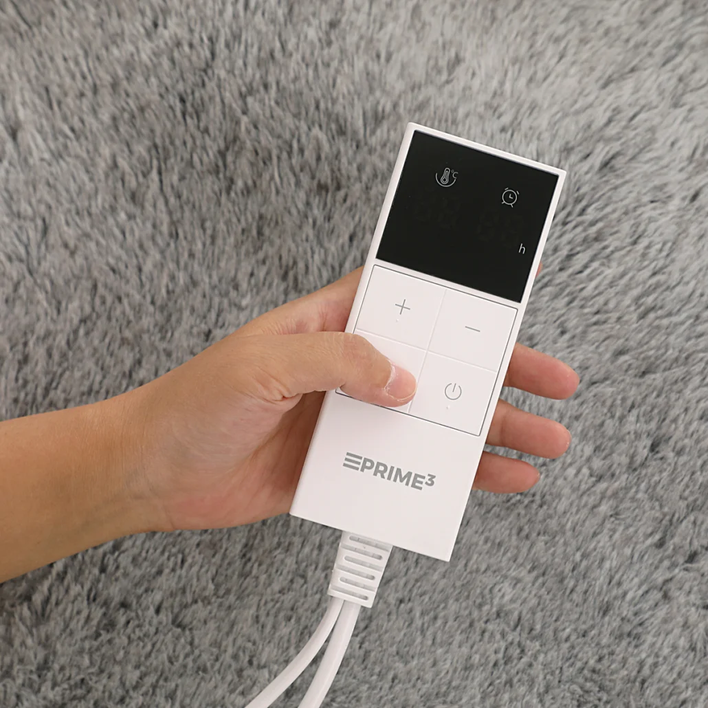 Ręka trzyma biały pilot z przyciskami i ekranem. Pilot jest podłączony do białego kabla. Tło jest szare.