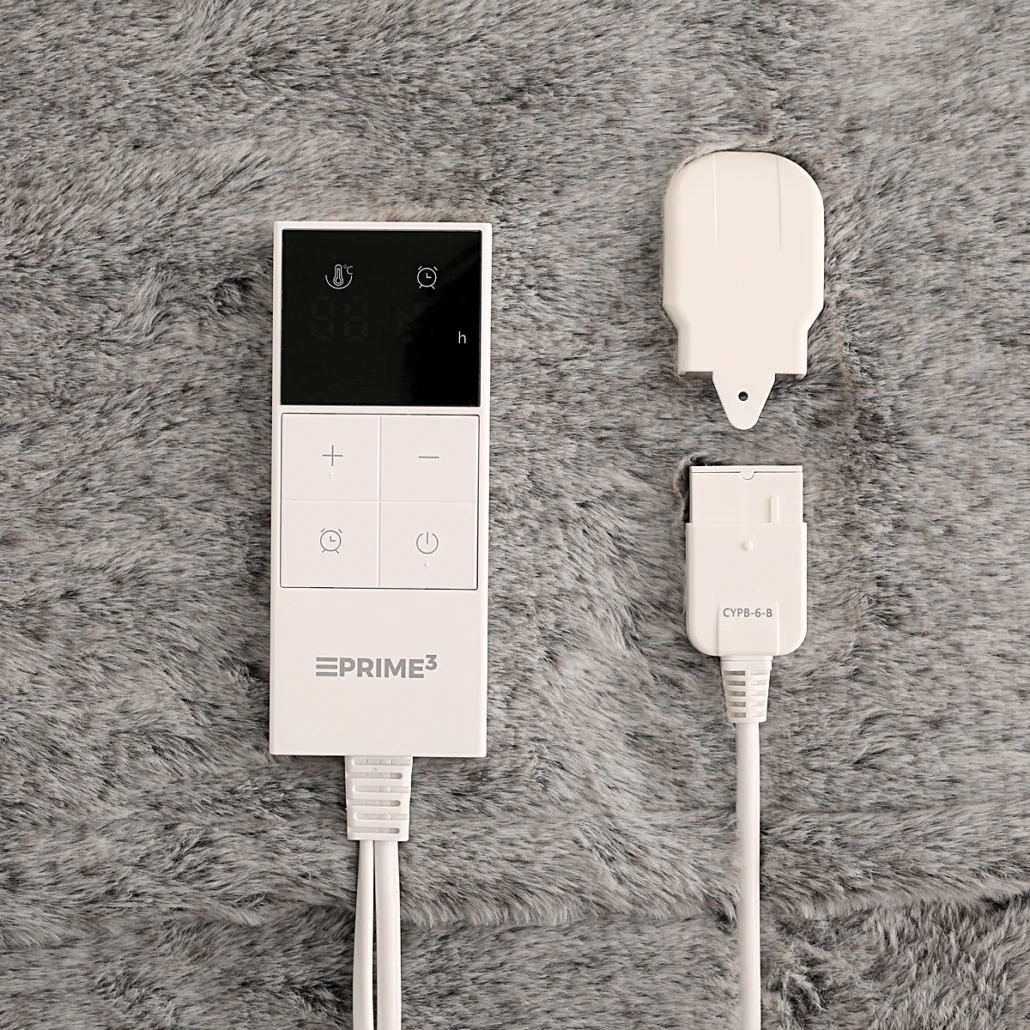 Biały kontroler i wtyczka do koca elektrycznego na szarym sztucznym futrze. Kontroler posiada przyciski i wyświetlacz. Wtyczka oznaczona CYPB-6-B.
