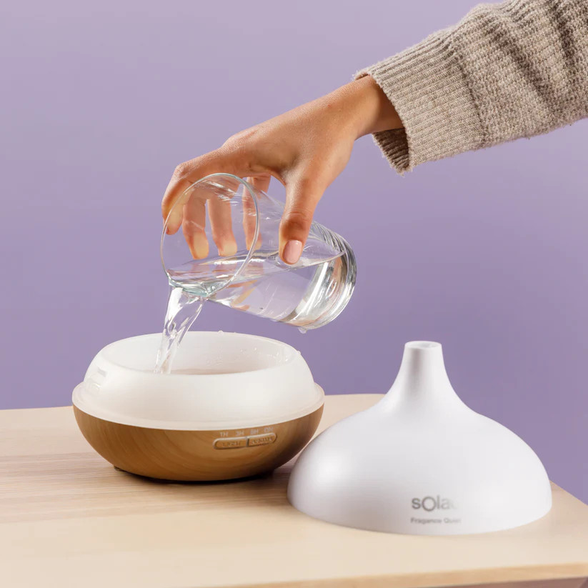 Ręka wlewa wodę ze szklanki do dyfuzora. Dyfuzor znajduje się obok białej pokrywy, na drewnianej powierzchni.