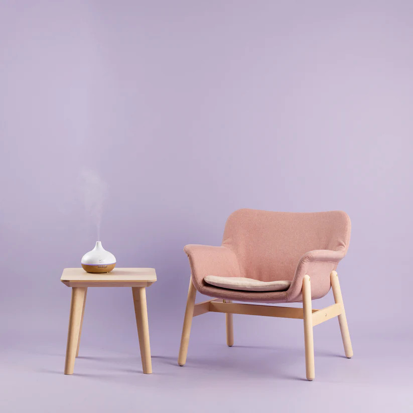 Różowe krzesło i mały drewniany stolik z dyfuzorem zapachu na fioletowym tle.