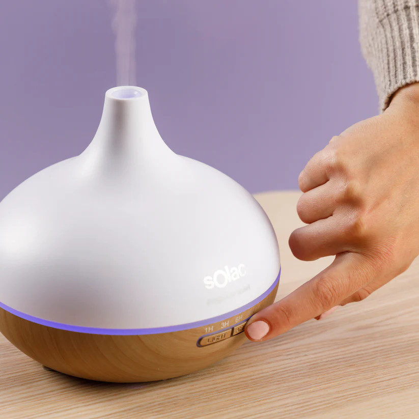 Biały dyfuzor Solac stoi na drewnianym stole. Ręka naciska przycisk. Para wydobywa się z góry. Fioletowa ściana w tle.