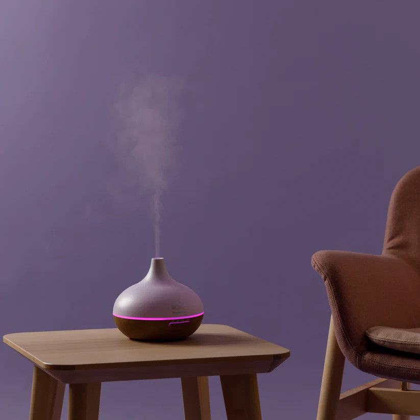 Biały i brązowy dyfuzor zapachów stoi na drewnianym stole. Krzesło po prawej stronie. Fioletowe tło.