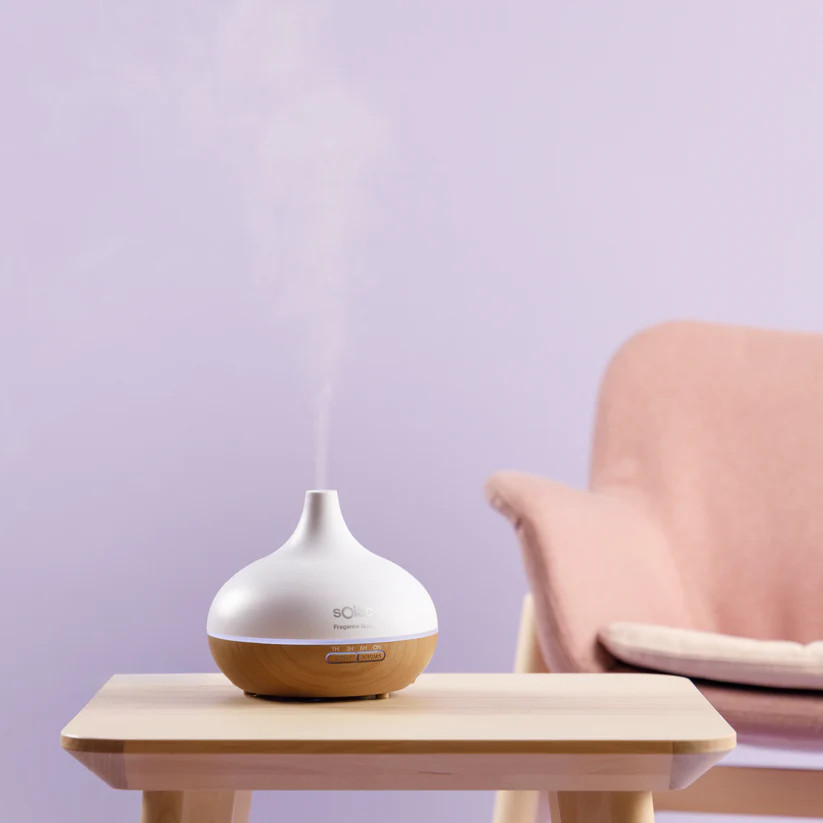 Biały i drewniany dyfuzor olejków eterycznych stoi na drewnianym stole. Różowe krzesło w tle na fioletowej ścianie.