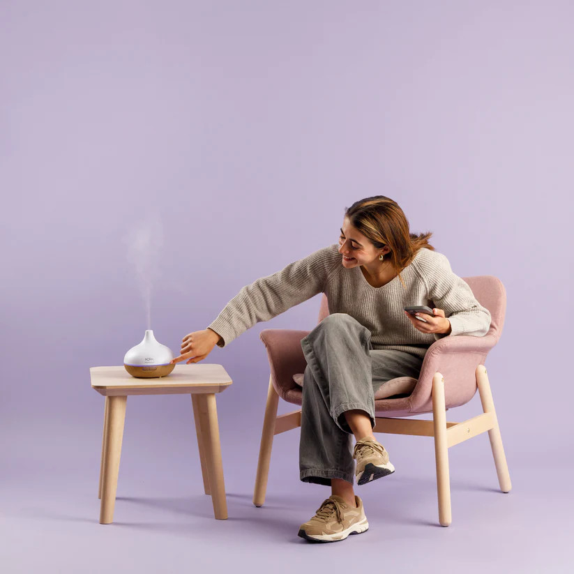 Kobieta siedzi na różowym krześle, dotykając białego dyfuzora na stoliku. Trzyma telefon. Tło jest fioletowe.