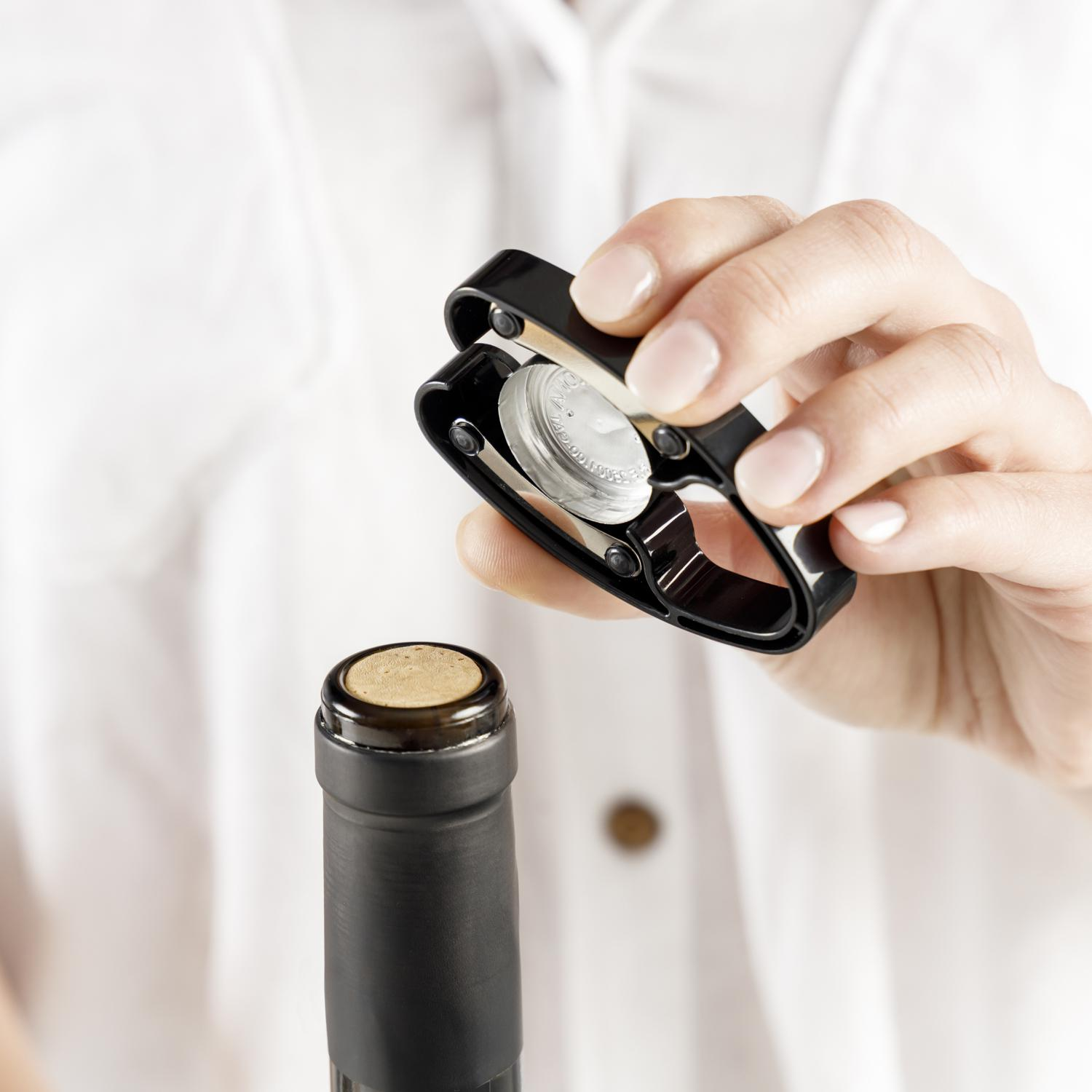 Ręka trzyma czarny otwieracz do wina nad butelką wina. Butelka ma korek i czarną etykietę. Biała koszula w tle.