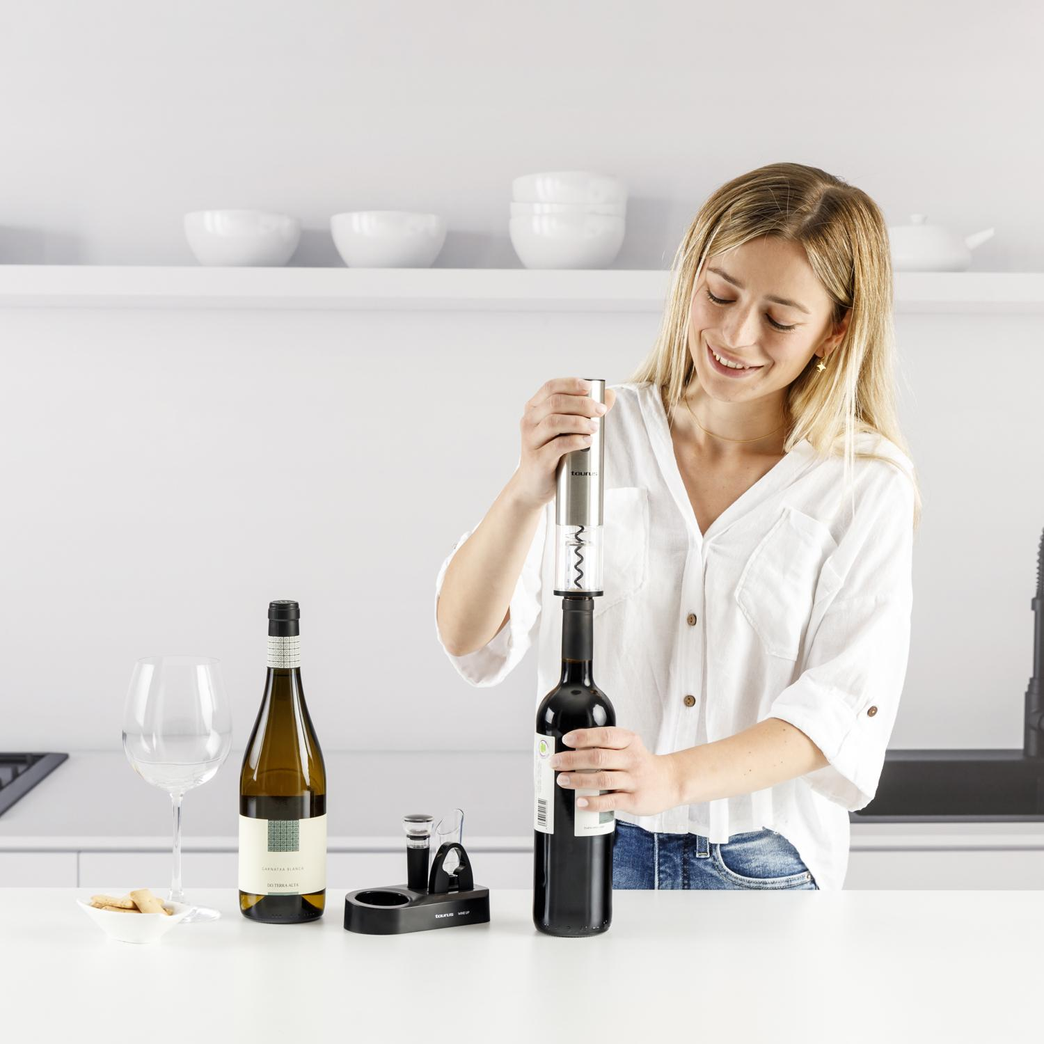 Kobieta otwiera butelkę wina korkociągiem. Inne elementy: kieliszek, butelka i przekąski.