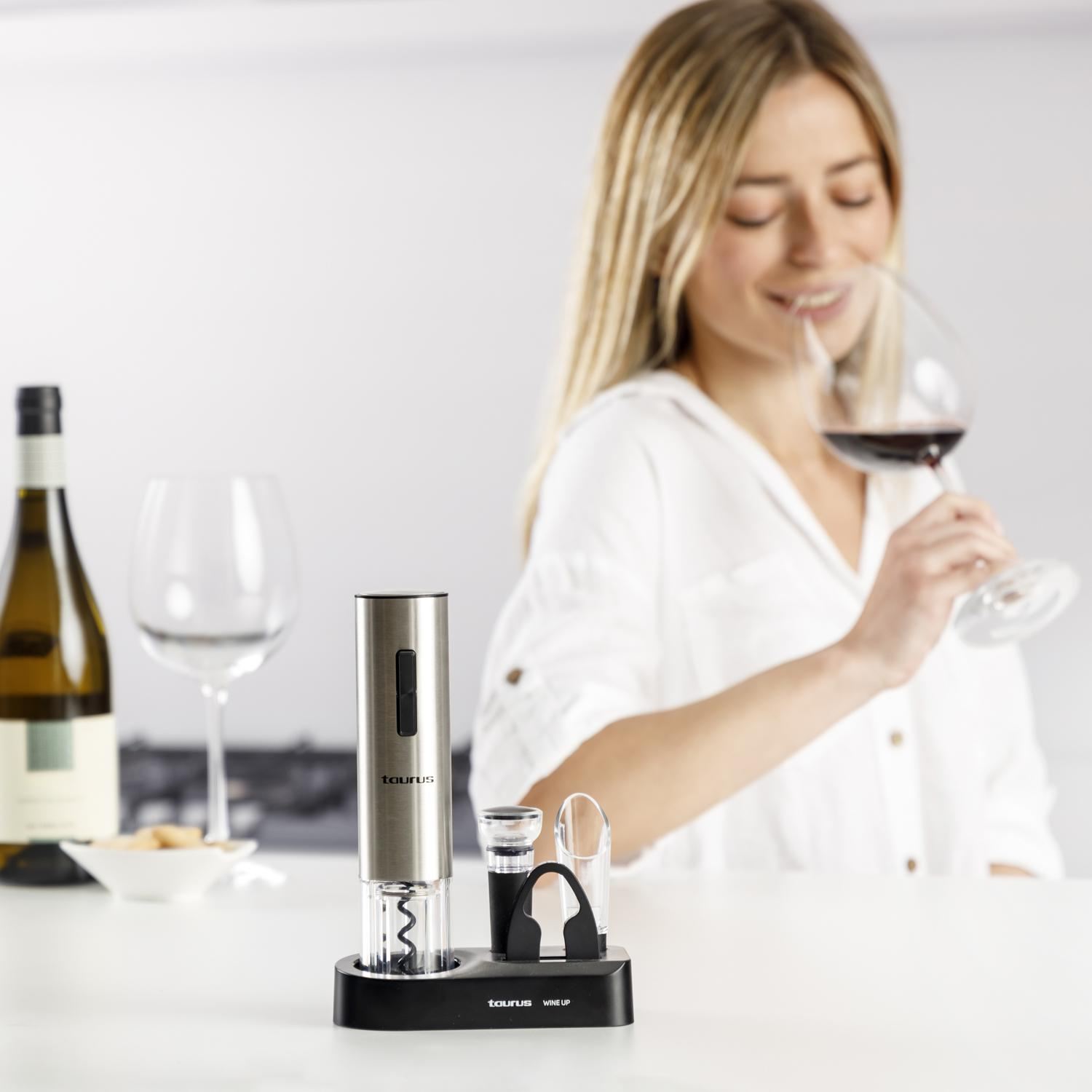 Kobieta wącha wino. Otwieracz do wina, butelka i szklanki na białym stole. Kobieta ma na sobie białą koszulę.