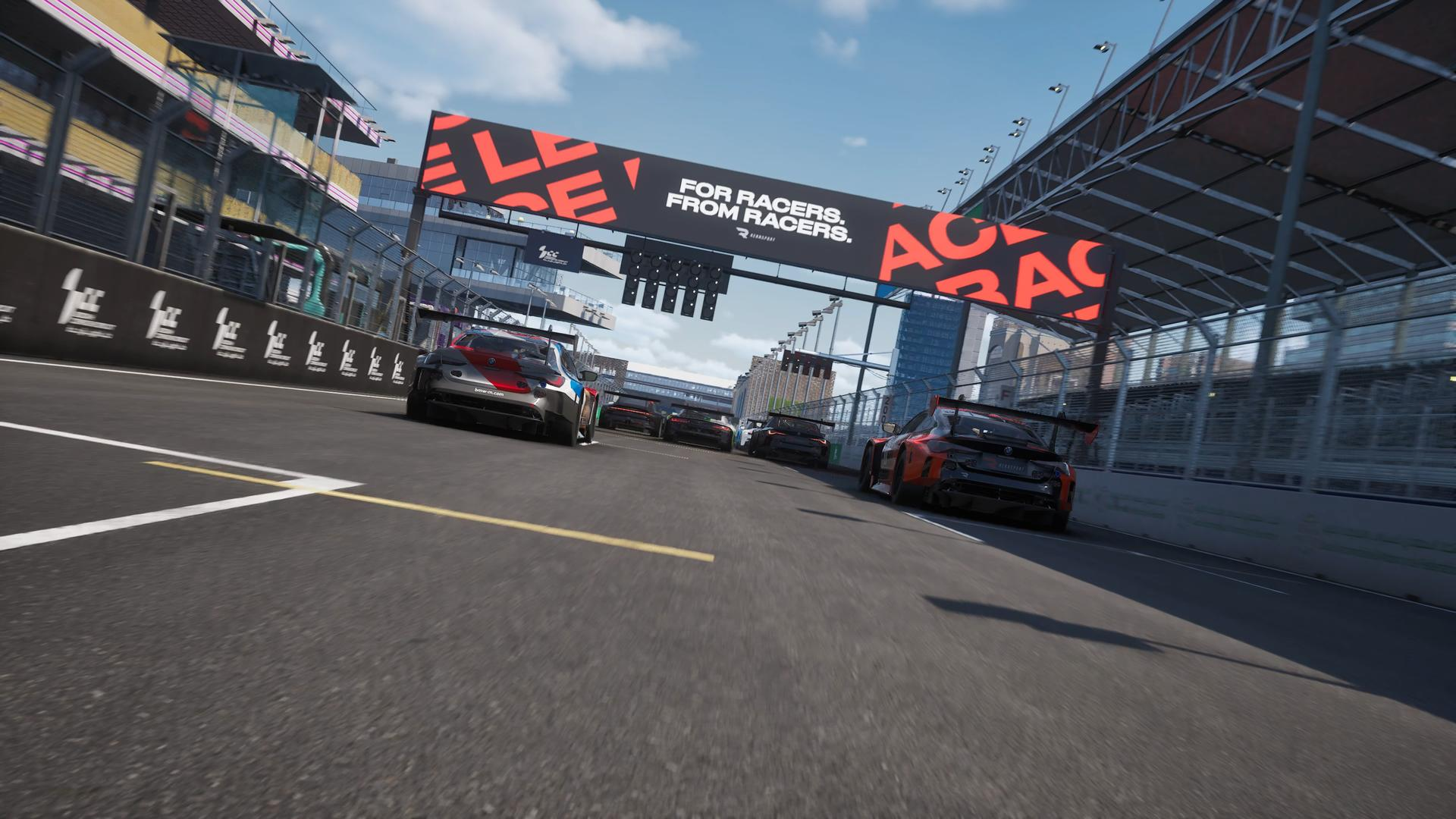 Rennwagen rasen auf einer Rennstrecke, mit einem Banner mit der Aufschrift 'For Racers. From Racers'. Gebäude und Himmel im Hintergrund.