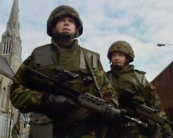 Zwei Soldaten in Tarnuniformen mit Gewehren stehen vor einer Kirche. Der Himmel ist bedeckt.