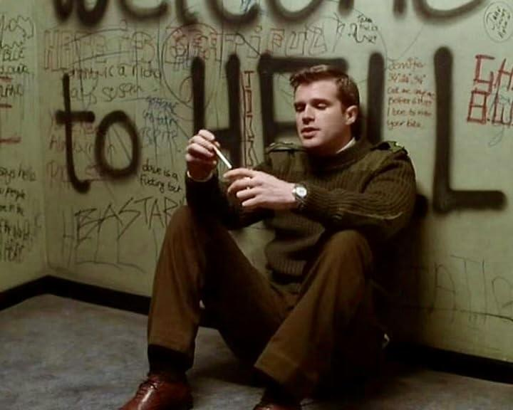 Ein Mann in grünem Pullover und brauner Hose sitzt an einer Wand mit Graffiti und hält ein kleines Objekt. An der Wand steht "WELCOME TO HELL".