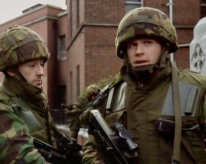 Zwei Soldaten in Tarnhelmen und Jacken mit Gewehren, im Hintergrund ein Gebäude. Andere Soldaten sind unscharf.
