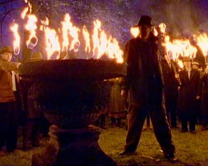 Ein Mann mit Hut steht vor einer großen Urne mit Feuer. Im Hintergrund halten Menschen Fackeln. Die Szene ist dunkel mit Feuerlicht.