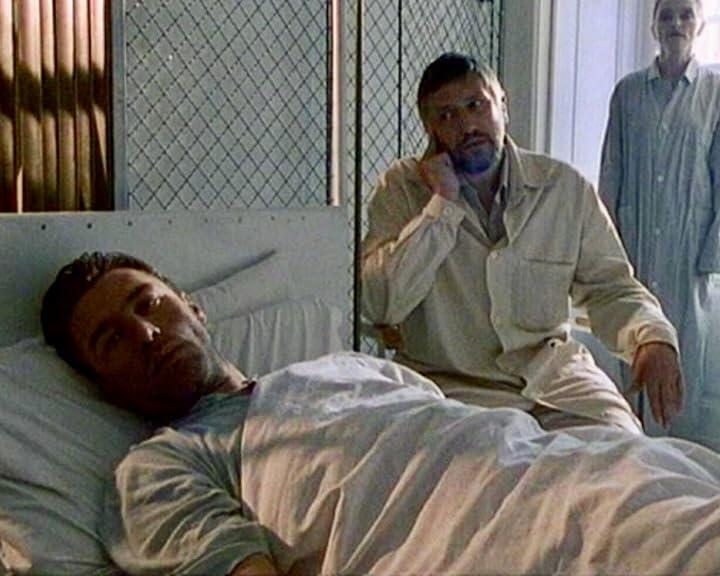 Ein Mann liegt im Krankenhausbett, ein anderer Mann sitzt daneben am Telefon, eine Frau steht im Hintergrund.