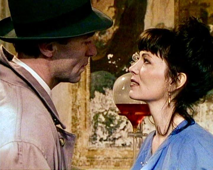 Ein Mann mit Hut und Mantel blickt eine Frau mit dunklen Haaren und blauem Oberteil an. Eine Laborflasche steht zwischen ihnen.