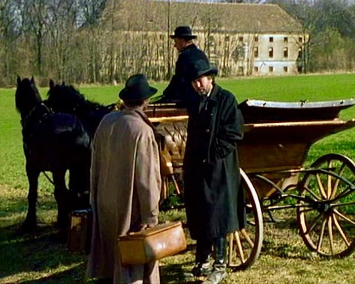 Männer mit Hüten an einer Pferdekutsche. Einer hält einen Koffer. Im Hintergrund ein großes Gebäude. Grünes Gras und Bäume sind sichtbar.