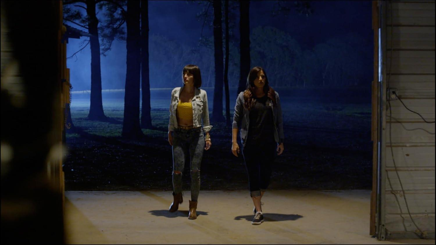 Zwei Frauen stehen in einer Garagenöffnung und blicken auf einen dunklen Wald. Eine trägt ein gelbes Oberteil und Jeans, die andere ein schwarzes Oberteil und Jeans.
