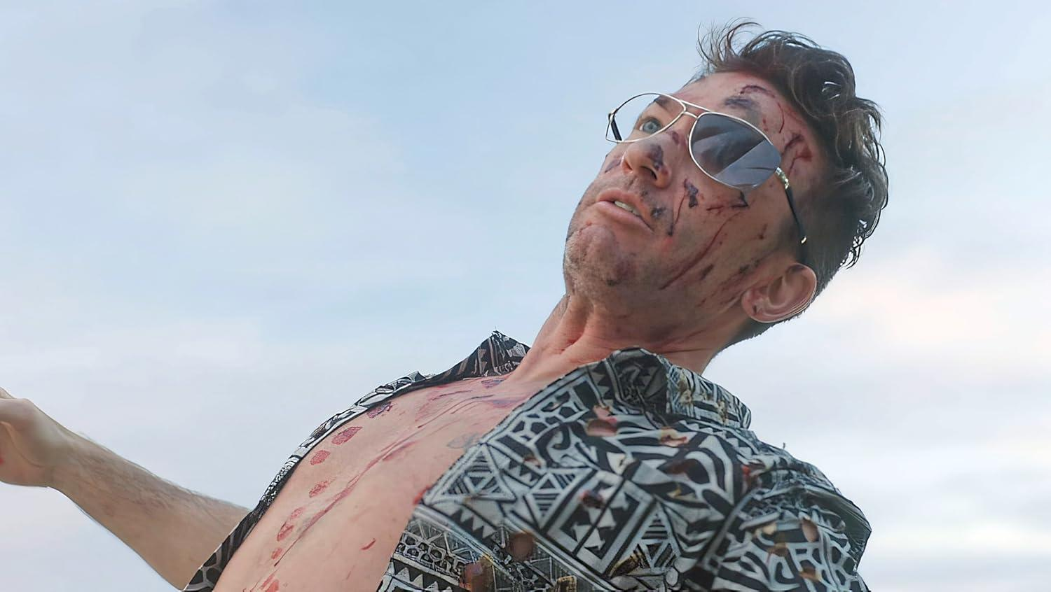 Ein Mann mit Sonnenbrille und gemustertem Hemd blickt nach oben. Sein Gesicht und seine Brust haben Schnitte und Blut, vor einem bewölkten Himmel.