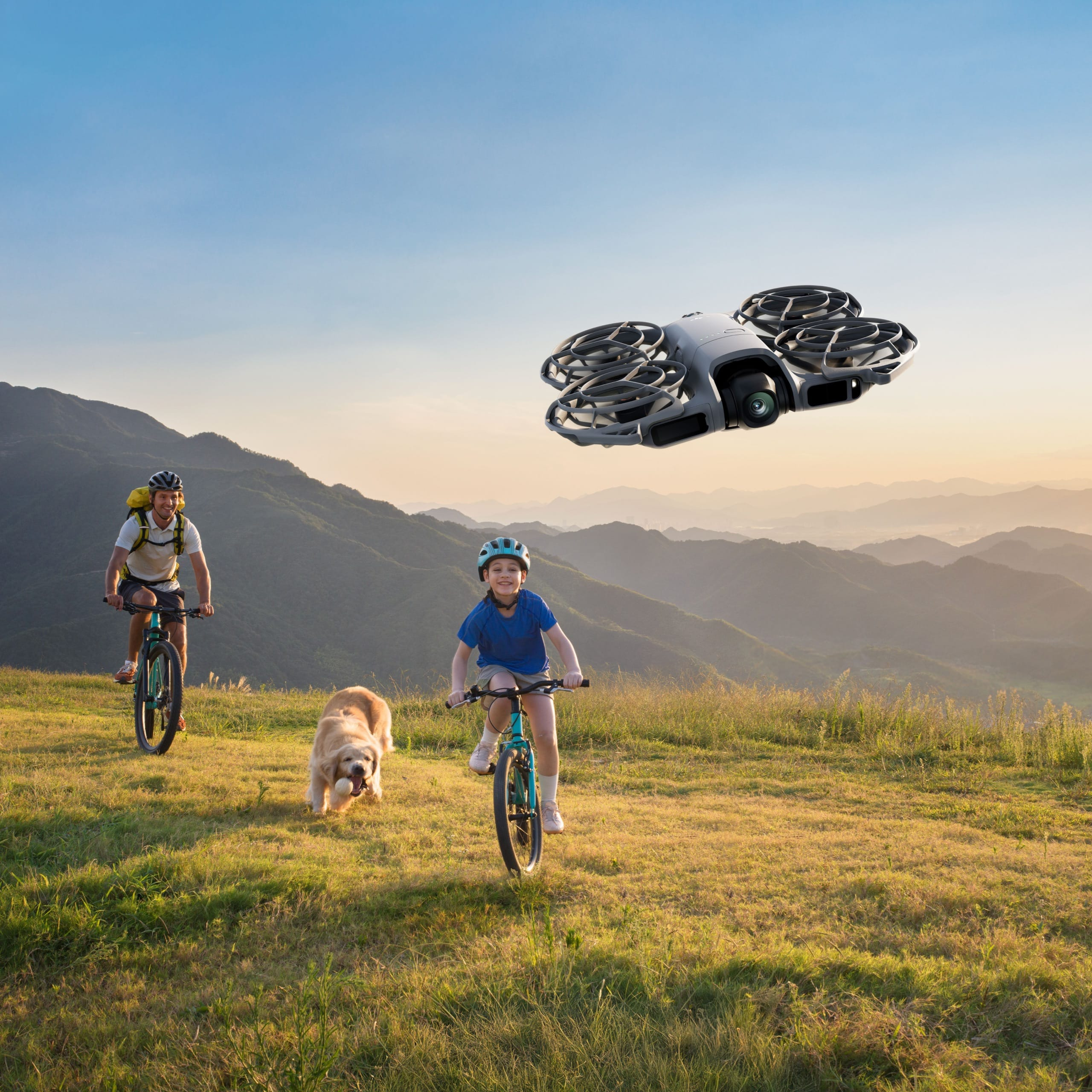 Des personnes font du vélo sur une colline herbeuse ; un drone vole au-dessus. Un chien court à côté. Montagnes en arrière-plan et ciel bleu.