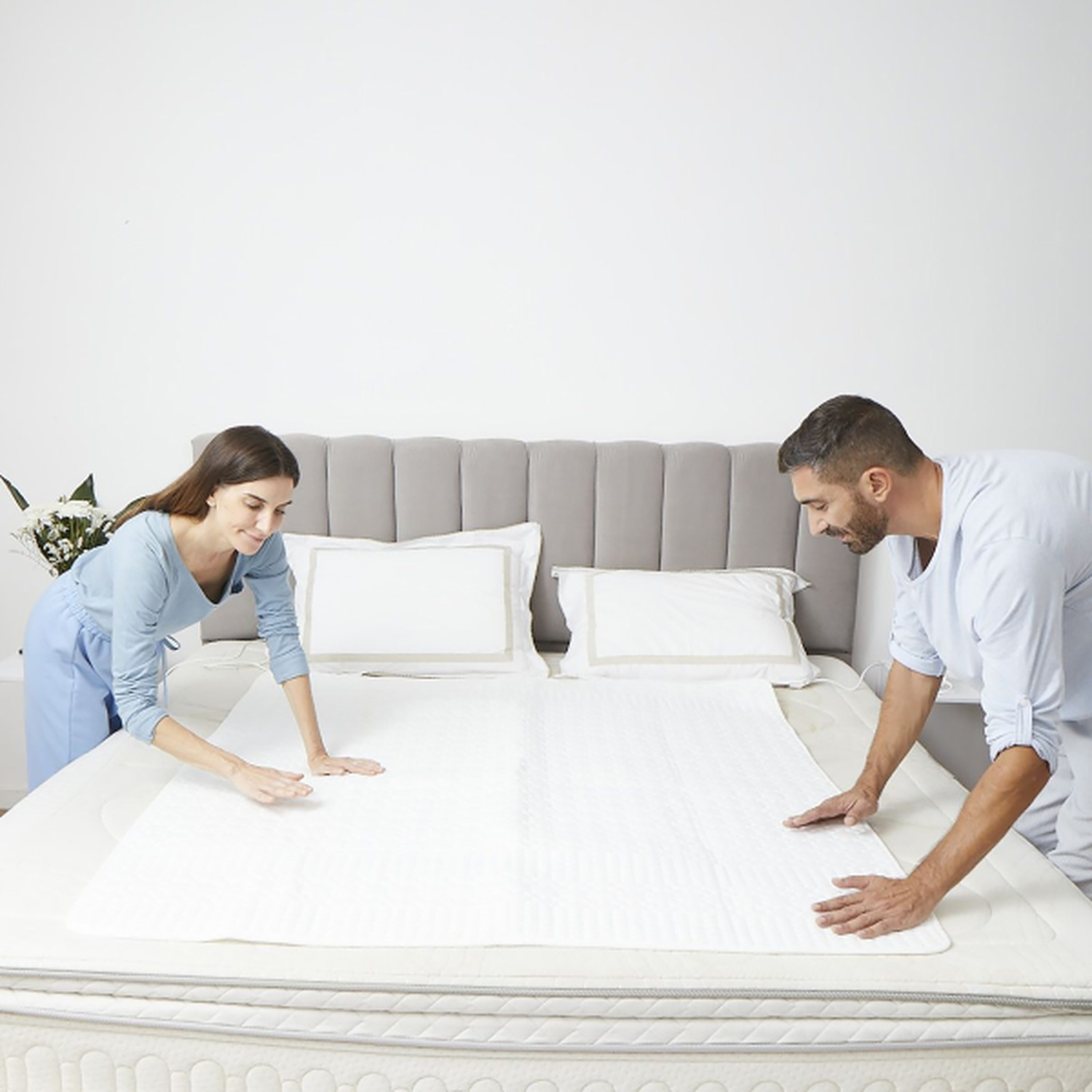 Un homme et une femme mettent un coussin blanc sur un lit. Le lit a des oreillers blancs et une tête de lit grise. Ils sourient tous les deux.