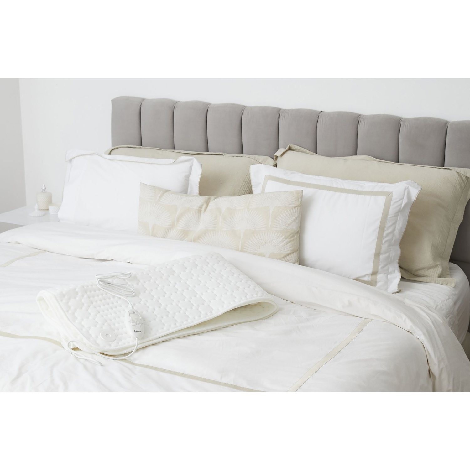 Un lit avec de la literie blanche, des oreillers et une tête de lit grise. Un coussin chauffant blanc est sur le lit. Une table de chevet à gauche.