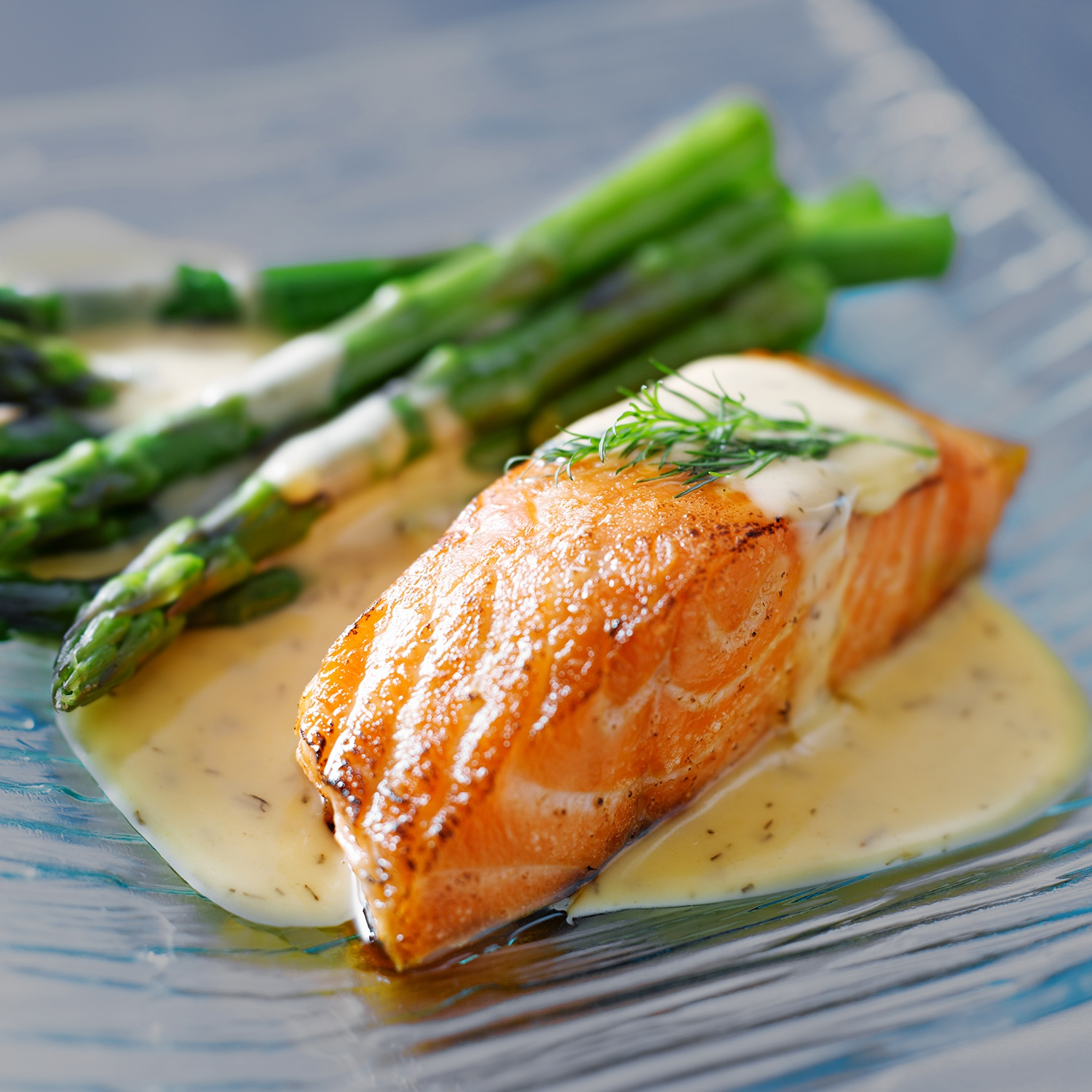 Salmone grigliato con asparagi e salsa cremosa su piatto di vetro. Asparagi verdi, salmone arancione e salsa gialla.