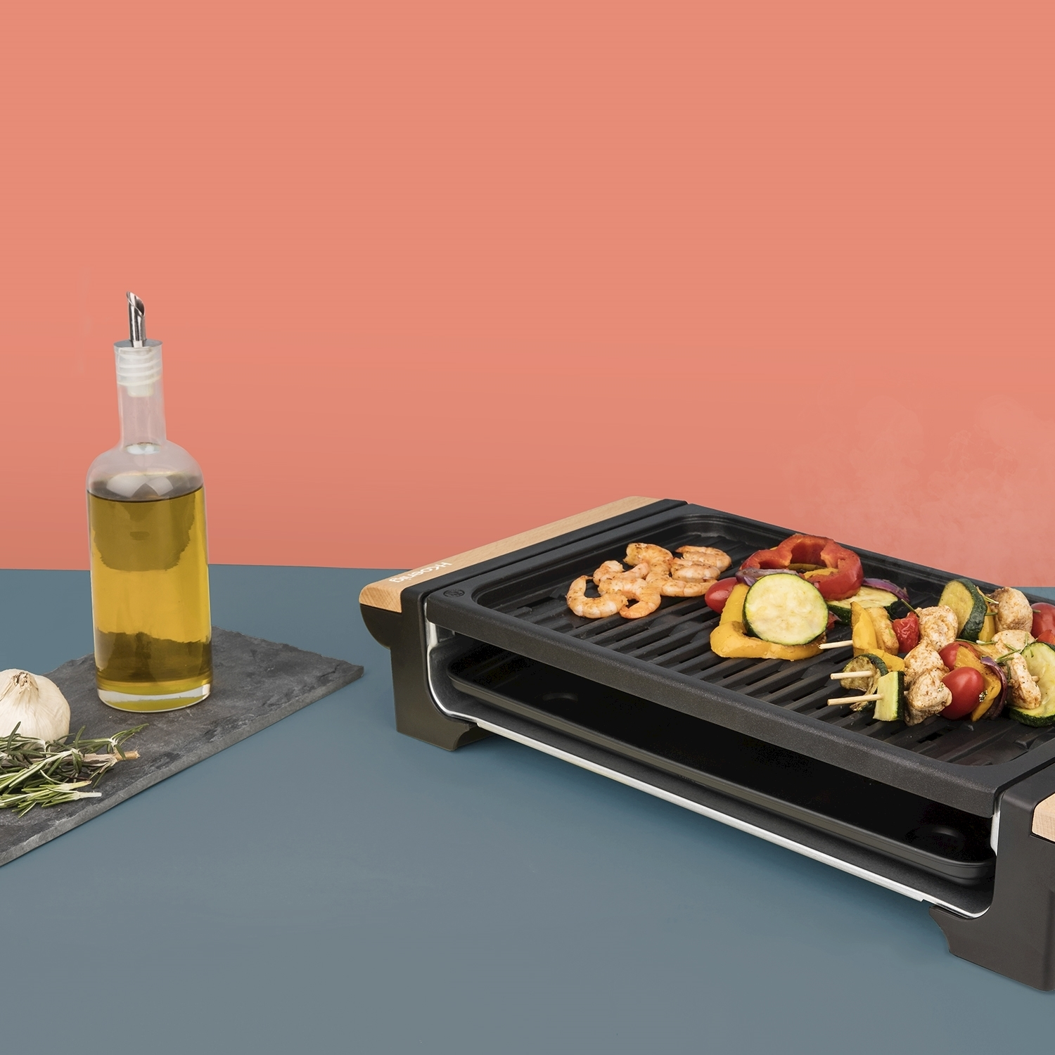 Griglia elettrica con spiedini di gamberi e verdure. Bottiglia di olio d'oliva e aglio a sinistra. Sfondo arancione e blu.