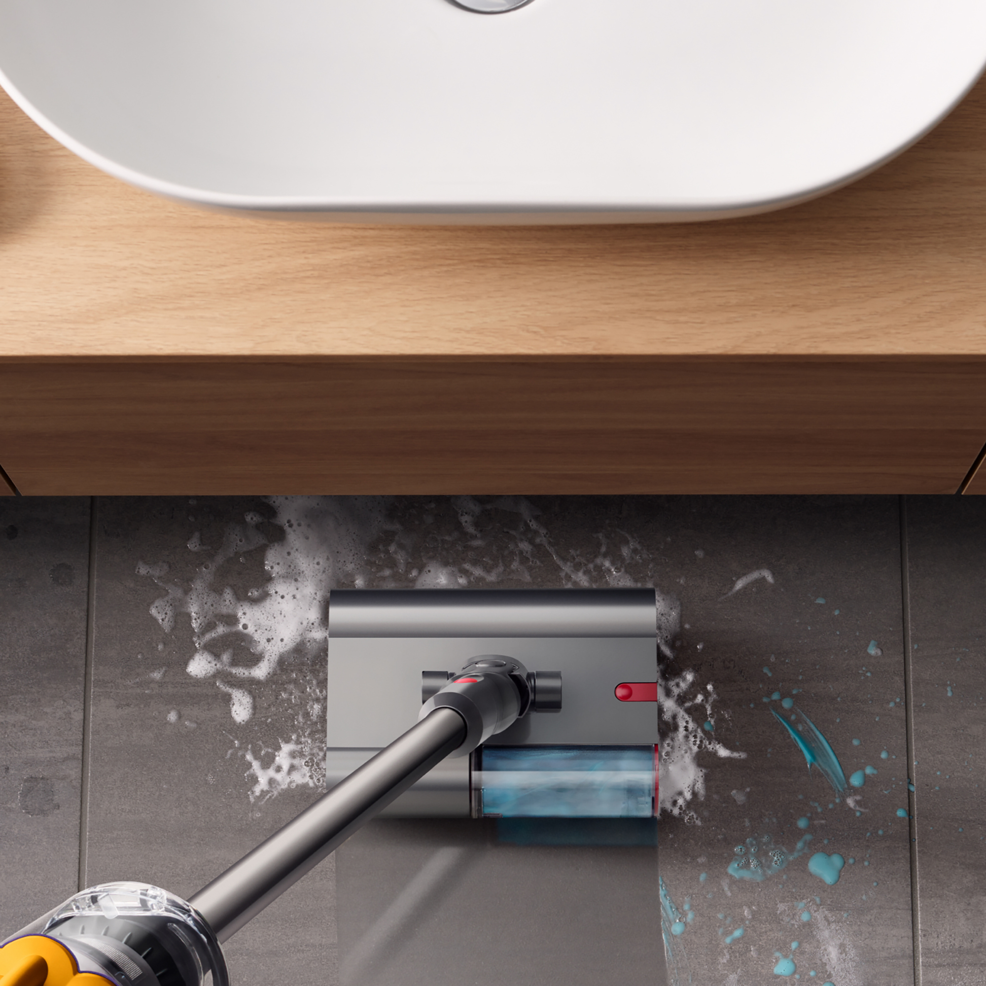 Szary odkurzacz czyści kafelkową podłogę mydłem. Biała umywalka i drewniana szafka w tle.